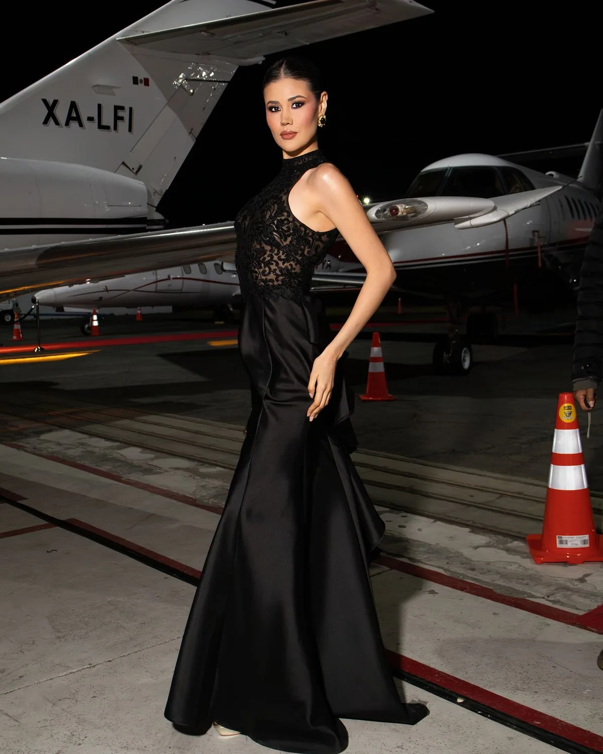 Woman in a black evening gown posing beside a private jet at night