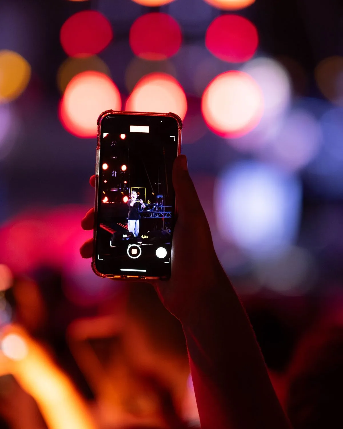 Hand holding up a phone to film a concert, stage lights blurred behind
